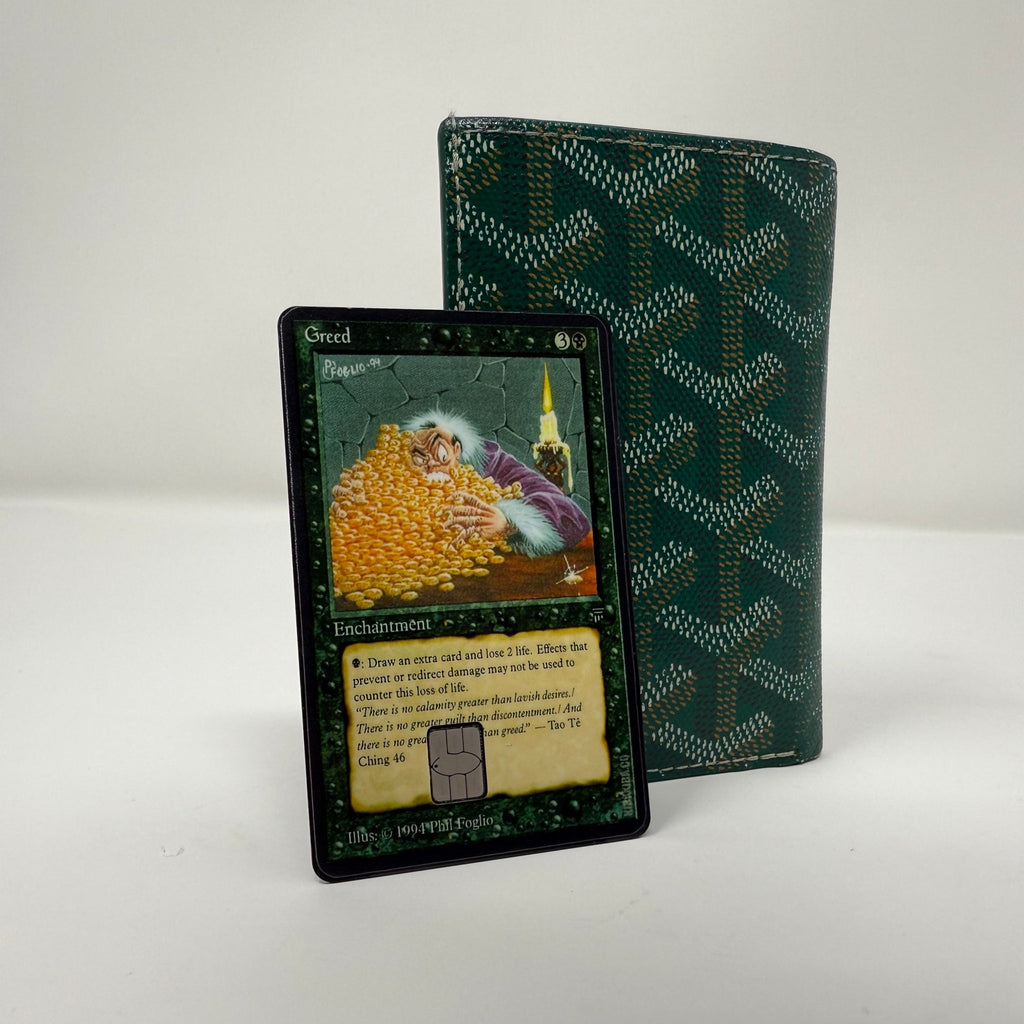 Single Greed Magic: The Gathering credit card sticker displayed at angle against green patterned wallet, showcasing detailed enchantment artwork with figure surrounded by golden coins and classic MTG card layout from Kibakiba.co